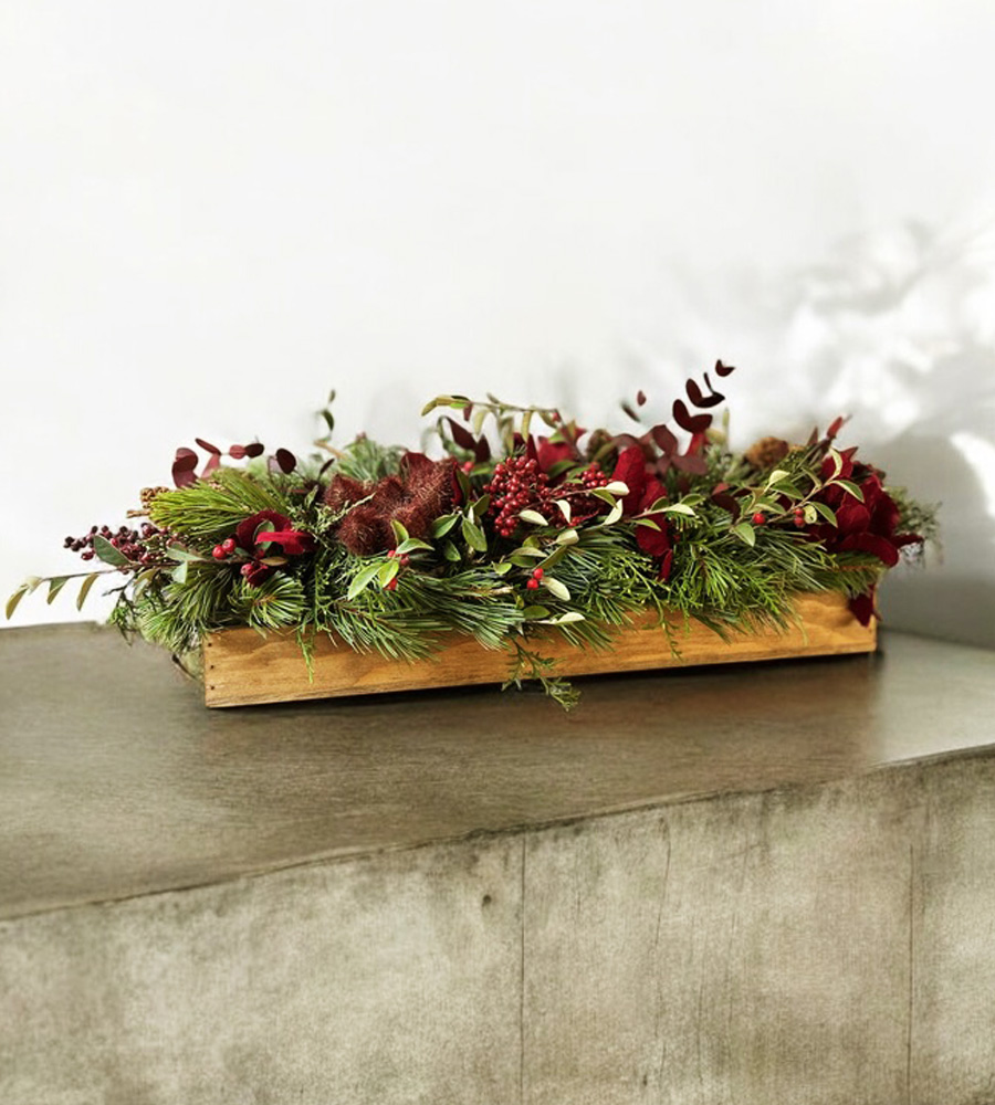 Centro de mesa navideño con follajes verdes, acentos rojos y base de madera